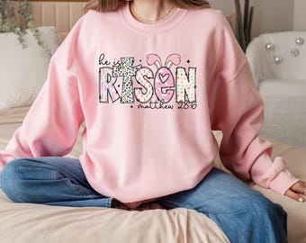 May include: A pink sweatshirt with the words "He is Risen" in colourful patterned letters. The text "Matthew 28:6" is written below the main text.