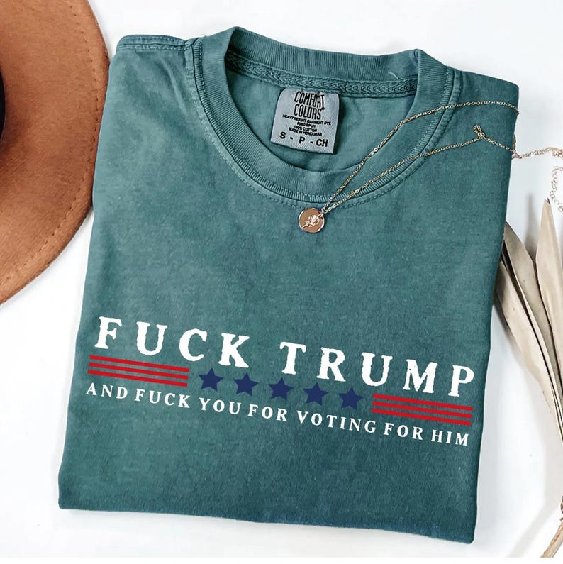 A green t-shirt with the text "FUCK TRUST" printed on it, along with the text "AND FUCK YOU FOR VOTING FOR HIM" below it. The t-shirt is laid out on a white surface, with a brown hat and some feathers visible in the background.