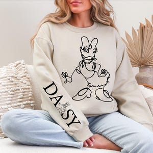 K&ouml;nnte beinhalten: Beiges Sweatshirt mit einer schwarzen Strichzeichnung von Daisy Duck. Auf dem linken &Auml;rmel steht das Wort "DAISY" in Schwarz, zusammen mit einer kleinen Minnie-Maus-Illustration. Das Sweatshirt wird mit hellblauen Jeans getragen.