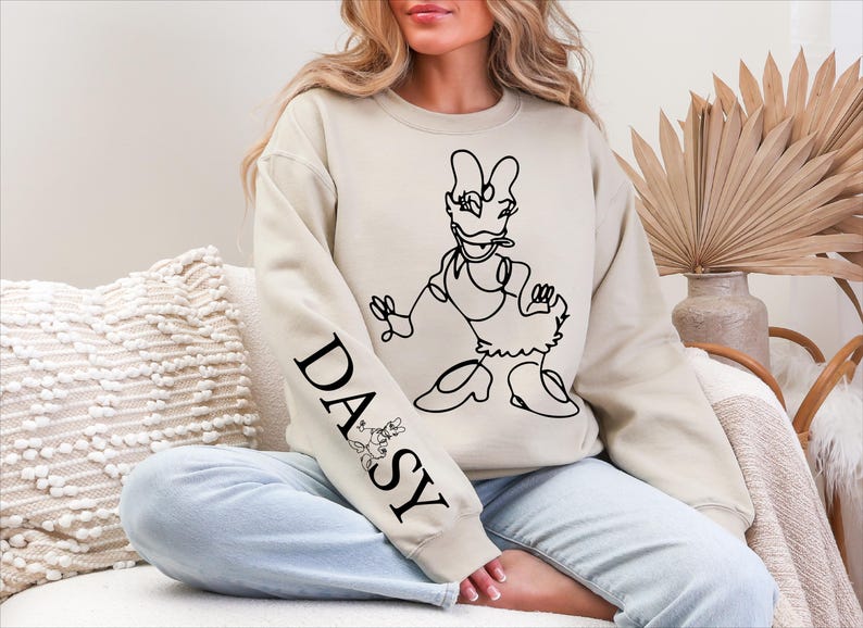K&ouml;nnte beinhalten: Beiges Sweatshirt mit einer schwarzen Strichzeichnung von Daisy Duck. Auf dem linken &Auml;rmel steht das Wort "DAISY" in Schwarz, zusammen mit einer kleinen Minnie-Maus-Illustration. Das Sweatshirt wird mit hellblauen Jeans getragen.