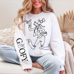 K&ouml;nnte beinhalten: Wei&szlig;es Sweatshirt mit einer schwarzen Strichzeichnung von Goofy und dem Wort "GOOFY" vertikal auf dem linken &Auml;rmel aufgedruckt. Das Design umfasst Goofys Hut, Handschuhe und Outfit. Ein lustiges, l&auml;ssiges Kleidungsst&uuml;ck.