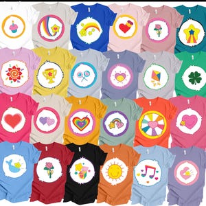 May include: A collection of colorful t-shirts featuring various cartoon-style designs. Each shirt has a different graphic in a white circle, including hearts, rainbows, stars, and flowers. The shirts come in a variety of colors, such as blue, pink, yellow, and green.