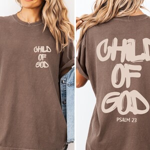 a woman wearing a brown t-shirt with the text "CHILD OF GOD" printed on it.