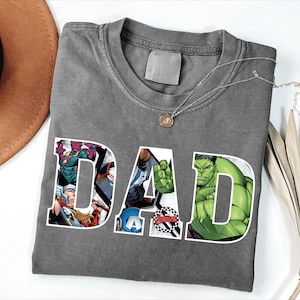 May include: Gray t-shirt with the word "DAD" in white block letters. The letters are filled with images of Marvel comic book characters, including Hulk. A silver necklace with a pendant is draped across the shirt.