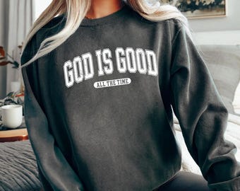 May include: Dark gray sweatshirt with the text "GOD IS GOOD" in white, arched across the chest, with "ALL THE TIME" below. The sweatshirt has long sleeves and a crew neck. The garment is a solid color.