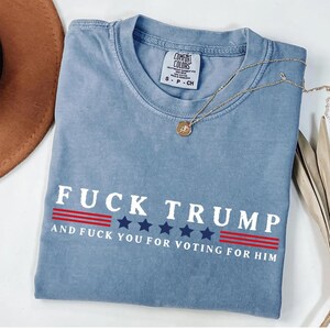 A blue t-shirt with the text "FUCK TRUST" printed on it, along with the text "AND FUCK YOU FOR VOTING FOR HIM" below it. The t-shirt is laid out on a white surface, with a brown hat and some leaves visible in the background.