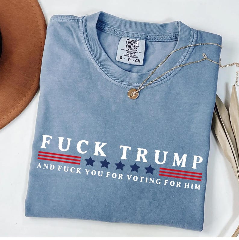 A blue t-shirt with the text "FUCK TRUST" printed on it, along with the text "AND FUCK YOU FOR VOTING FOR HIM" below it. The t-shirt is laid out on a white surface, with a brown hat and some leaves visible in the background.
