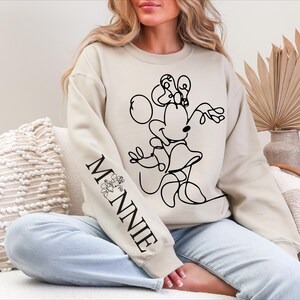 K&ouml;nnte beinhalten: Beiges Sweatshirt mit einer schwarzen Strichzeichnung von Minnie Mouse. Der &Auml;rmel zeigt das Wort "MINNIE" in Schwarz mit einem floralen Design. Ein l&auml;ssiger, bequemer Kleidungsstil.