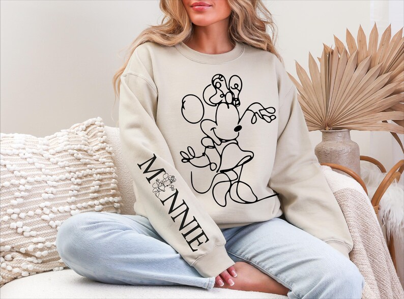 K&ouml;nnte beinhalten: Beiges Sweatshirt mit einer schwarzen Strichzeichnung von Minnie Mouse. Der &Auml;rmel zeigt das Wort "MINNIE" in Schwarz mit einem floralen Design. Ein l&auml;ssiger, bequemer Kleidungsstil.