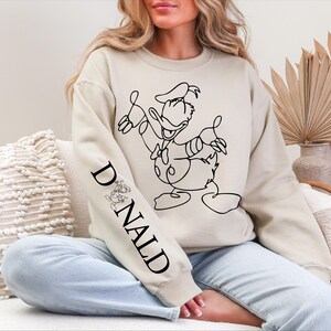 K&ouml;nnte beinhalten: Beiges Sweatshirt mit einer schwarzen Strichzeichnung von Donald Duck. Der &Auml;rmel zeigt das Wort "DONALD" in Schwarz, begleitet von einer kleinen Donald Duck-Illustration. Ein l&auml;ssiger, bequemer Stil.