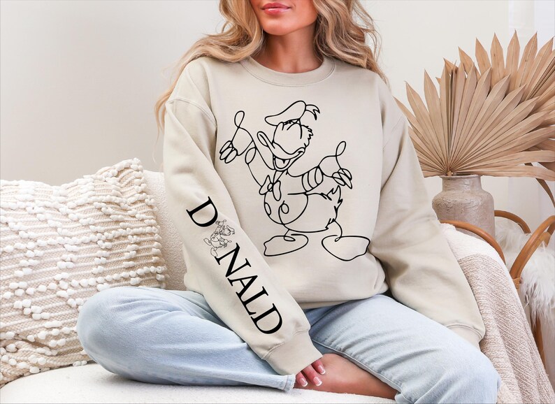 K&ouml;nnte beinhalten: Beiges Sweatshirt mit einer schwarzen Strichzeichnung von Donald Duck. Der &Auml;rmel zeigt das Wort "DONALD" in Schwarz, begleitet von einer kleinen Donald Duck-Illustration. Ein l&auml;ssiger, bequemer Stil.