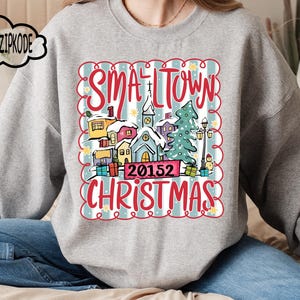 May include: A grey sweatshirt with a festive graphic. The design features the words "Small Town Christmas" in red, along with a winter village scene, a church, houses, and a Christmas tree. The zip code "20152" is also displayed.