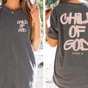 a woman wearing a gray t-shirt with the text "CHILD OF GOD" printed on it. The back of the t-shirt also displays the text "PSALM 23" along with a graphic of a Bible verse.