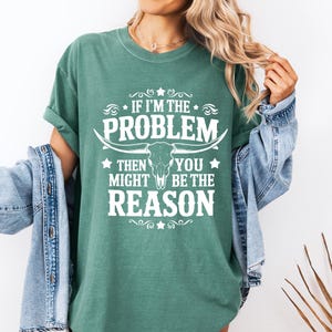 May include: A green t-shirt with the text "IF I'M THE PROBLEM THEN YOU MIGHT BE THE REASON" in white. The design includes a bull skull and decorative elements. The shirt is part of a casual wear collection.