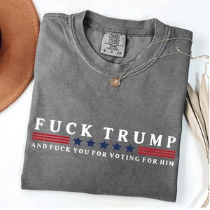 A gray t-shirt with the text "FUCK TRUST" printed on it, along with the text "AND FUCK YOU FOR VOTING FOR HIM" below it. The t-shirt is laid out on a white surface, with a brown hat and some beige fabric visible in the background.