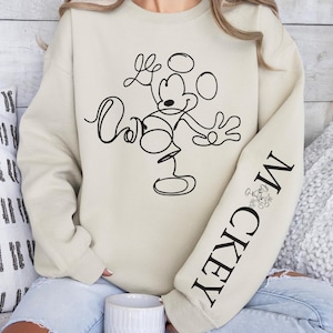 May include: Cream-colored sweatshirt featuring a black line drawing of Mickey Mouse. The sleeve displays the word "Mickey" in black, along with a small Mickey Mouse graphic. The sweatshirt is part of a Disney-themed apparel collection.