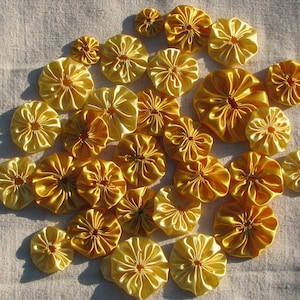 May include: A collection of yellow fabric rosettes, arranged in a circular pattern on a white fabric background. The rosettes are made of satin and have a ruffled appearance.