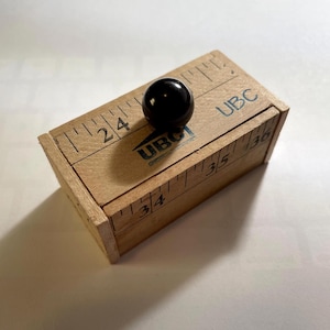 May include: A rectangular wooden block with a smooth, light brown finish, featuring black printed ruler markings and the text UBC. A small, glossy black spherical knob is attached to the top surface, which displays measurements ranging from 24 to 36 inches.