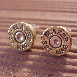 May include: A pair of brass-colored stud earrings. Each earring is shaped like a bullet casing, with the text "BLAZER MAG .357" engraved around the edge. The earrings have a metallic silver center and are set against a brown background.