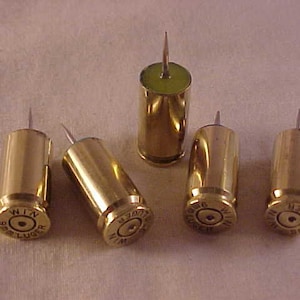 May include: Five gold-colored bullet casings with a small pin attached to the top. Each casing has a different inscription on the side, including "WIN 9mm LUGER" and "9mm Luger".