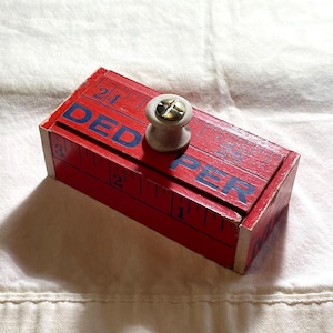 May include: A rectangular red wooden box with a white spool-shaped knob on top. The box features blue printed text that reads DED PER and black ruler markings along the edges, including numbers 2, 3, 24, and 26, with a metallic screw in the center.