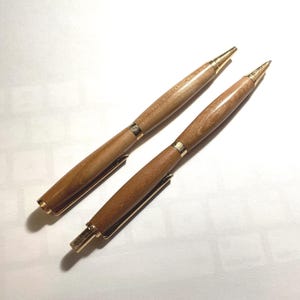 May include: Two pens with light brown wooden barrels and gold-colored metal accents. Each pen features a gold clip, a gold tip, and a gold push-button mechanism at the top. The wood grain is visible on the cylindrical bodies of both writing instruments.