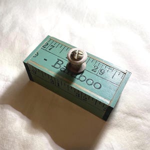 May include: A rectangular teal wooden box featuring a printed ruler pattern with black markings and numbers. The top surface displays the text "Baraboo" in black lettering. A small, round, light-colored wooden knob with a brass screw is centered.