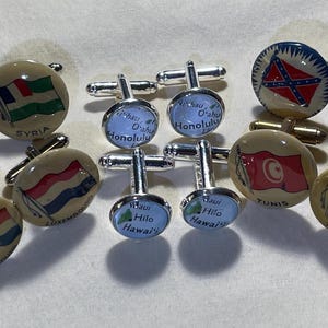 May include: A collection of cufflinks with various designs. Some feature flags of different countries, while others display the text "Honolulu" and "Hawaii". The cufflinks have a silver or gold finish and are set against a neutral background.