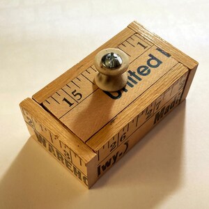 May include: A rectangular wooden box with a light brown finish, featuring printed ruler markings along the edges and the text United, WAREHO, and Madi. A small, light-colored wooden spool handle with a silver metal screw is attached to the top.