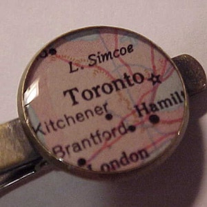 May include: A silver tie clip with a round glass cabochon featuring a map of Toronto, Ontario. The map shows the city's major roads and surrounding towns, including Kitchener, Brantford, Hamilton, and London.