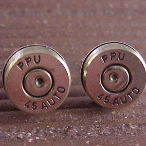 May include: A pair of metallic cufflinks, each resembling a .45 Auto bullet casing. The casings are a brassy gold color with the markings "PPU" and "45 AUTO" engraved on the face. The cufflinks have a shiny, polished finish.