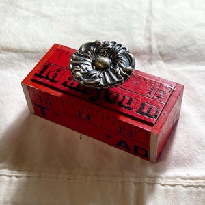 May include: A rectangular red wooden box featuring a printed measuring tape design with black markings and numbers. The lid is topped with a round, ornate, metallic silver-toned knob with intricate floral-inspired embossed detailing.