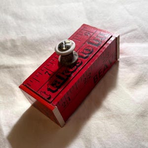 May include: A rectangular red wooden box with a white and metallic knob on top. The box features black printed ruler markings and the text "it takes to be" and "BEA" visible on the sides. The design mimics a measuring tape with clear inch increments.
