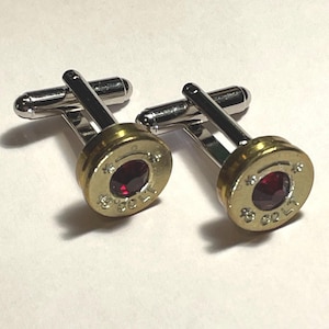 May include: A pair of cufflinks crafted from brass-colored bullet casings with silver-toned metal toggle backings. Each casing features a deep red circular gemstone set in the center and the text 45 COLT stamped into the metal surface.