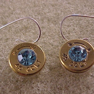May include: A pair of gold-colored earrings with blue rhinestones set in the center of each earring. The earrings are made from repurposed .45 Colt bullet casings.