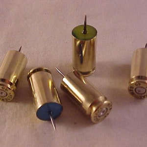 May include: Five brass push pins with sharp metal points. The push pins are made from spent bullet casings. One push pin has a green top, and another has a blue top. The casings are stamped with the number 45.