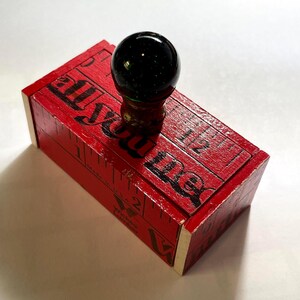 May include: A rectangular red wooden stamp with a black spherical handle. The top surface features black printed text that reads "all you me" and includes markings resembling a ruler with the text "Made in U.S.A." and "Wicked Lumber".