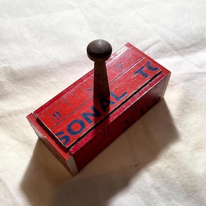 May include: A rectangular red wooden stamp with a dark brown wooden handle. The top surface features a printed ruler scale and the blue text SONAL TO. The stamp has a smooth finish and a block-like structure, designed for manual use with ink.