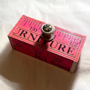 May include: A rectangular wooden box with a pink surface printed with black measuring tape markings and the text FURNITURE and 163 South. A small, light-colored circular knob with a central metal screw is attached to the top of the box.