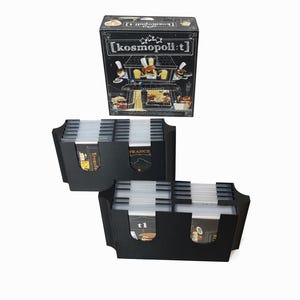 May include: A black board game box and two black card holders. The box has the text "[kosmopoli:t]" and a graphic of a restaurant. The card holders have clear plastic dividers and labels with the word "France".
