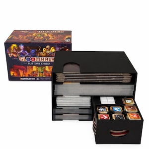 Op de afbeelding: Een zwarte bordspellen organizer met meerdere compartimenten, met kaarten, tegels en andere spelonderdelen. De doos ernaast toont de speltitel "Gloomhaven: Buttons & Bugs" met kleurrijke fantasy-kunst.