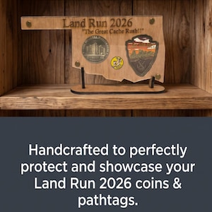 May include: A wooden display stand shaped like the state of Oklahoma, featuring the engraved text Land Run 2026 and The Great Cache Rush. The display holds a circular metallic coin, a small circular yellow tag, and a shield-shaped patch with a landscape.