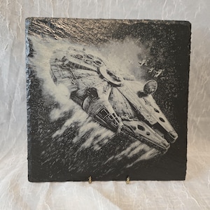 Könnte beinhalten: Eine quadratische schwarze Schieferplatte mit einer detaillierten, weiß lasergravierten Illustration des Millennium Falcon Raumschiffs im Flug. Das Kunstwerk zeigt das Schiff, wie es sich mit Bewegungslinien und kleineren Raumschiffen im Hintergrund durch den Weltraum bewegt.