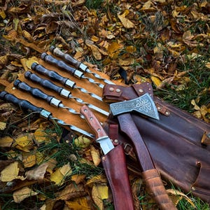 Puede incluir: Un juego de seis pinchos metálicos con mangos de madera, un cuchillo con funda de cuero y un hacha con funda de cuero y mango de madera. El juego está dispuesto sobre un lecho de hojas marrones.