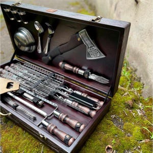 Puede incluir: Un juego de caja de madera con una variedad de herramientas para asar a la parrilla, incluyendo pinchos, una rejilla para asar, un peque&ntilde;o hacha y una esp&aacute;tula. La caja est&aacute; forrada con madera oscura y tiene un asa de cuero.