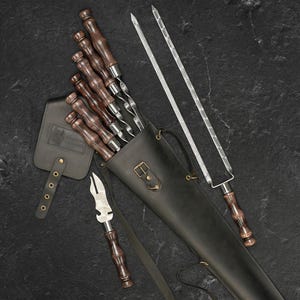 May include: A set of grilling skewers with dark wooden handles and stainless steel blades, stored in a black leather carrying case. Includes a leather tool holder and two long, flat skewers. The set is designed for outdoor cooking.