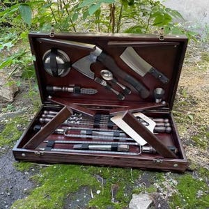 May include: A wooden box with a hinged lid, containing a set of grilling tools. The tools include skewers, a spatula, a knife, and a small axe. The box is lined with a dark brown fabric.
