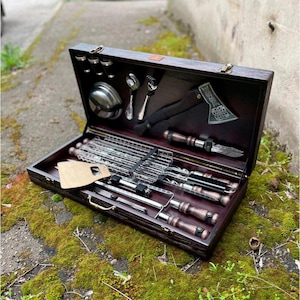 Puede incluir: Una caja de transporte de madera con un juego de herramientas para asar, que incluye pinchos, una rejilla para asar, una espátula y un pequeño hacha. La caja está forrada con fieltro marrón y tiene un cierre de latón.