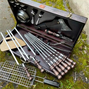 Puede incluir: Un juego de caja de madera con una variedad de herramientas para asar a la parrilla, incluyendo pinchos, una rejilla para asar, pinzas y un peque&ntilde;o hacha. La caja est&aacute; abierta y las herramientas est&aacute;n ordenadas cuidadosamente en el interior.