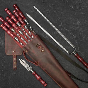 May include: A set of stainless steel skewers with red wooden handles, a carving fork, and a leather carrying case. The case is brown with a shoulder strap and a small leather pouch. The skewers are designed for grilling.